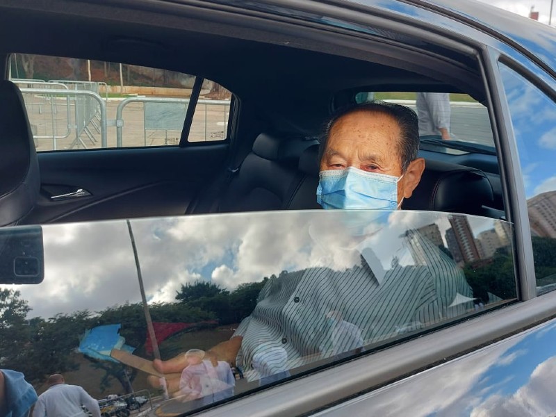 Senhor idoso chega no banco traseiro do carro para ser vacinado. Ele está de máscara e de camisa listrada
